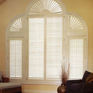 Plantation shutter grouping consisting of various shutter shapes covering an atrium window in the center with a complete arched shutter on the top with single shutter panels on the side windows with half arch shutters on top of them.