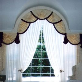 Beautiful arch window treatment featuring swags and jabots with tassel fringe, pinch pleated draperies styled in bishop sleeve, and sheer drapes.