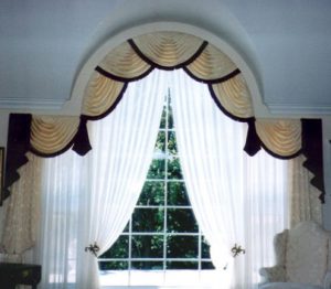 Beautiful arch window treatment featuring swags and jabots with tassel fringe, pinch pleated draperies styled in bishop sleeve, and sheer drapes.