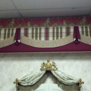 This top photo shows another example of a straight cornice withy flat swags and pelmets in coordinating fabrics and decorative trim. Below is a throw swag valance in a printed sheer fabric with bullion fringe on the inside edge mounted on three decorative hardware brackets mounted over top of an arched pair of plantation shutters. 
